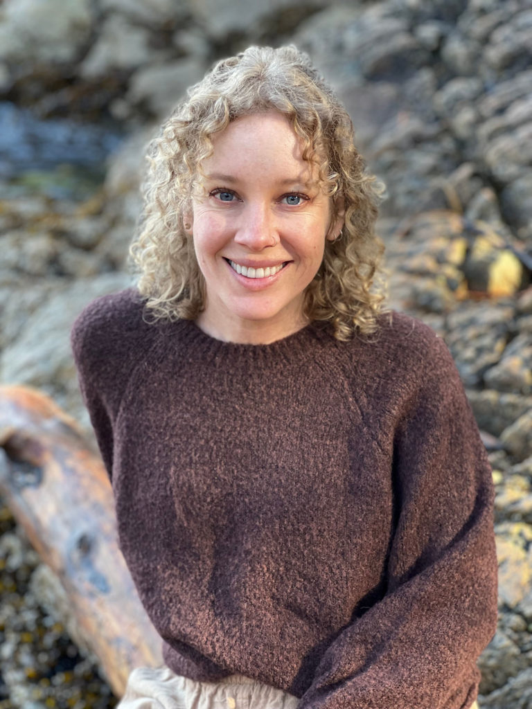 Shannon Tully white woman with blond curly hair. On a beach sitting on a log and looking at the camera and smiling.